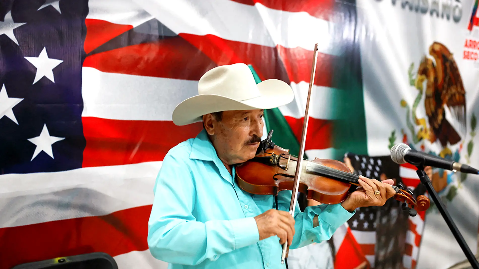 Hombre toca el violín y al fondo con banderas de México y Estados Unidos